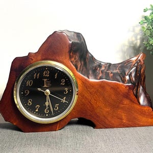 May include: A wooden desk clock with a black clock face and gold-colored trim. The clock is set in a uniquely shaped, polished wood base. The wood has a rich, reddish-brown color with darker, textured areas.
