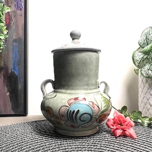 May include: A grey ceramic jar with a lid, decorated with colourful floral designs. The jar has two handles and a blue and white floral pattern on the front.