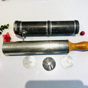 May include: A set of vintage cookie press tools, including a silver-colored cookie press with a wooden handle, a silver-colored tube, and three cookie cutters. The cookie cutters are shaped like a star, a snowflake, and a wavy line.