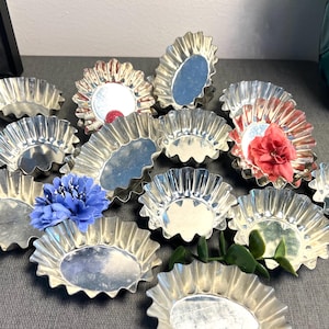 May include: A collection of small, fluted, silver-colored tart pans, some with colorful flower accents. The pans are arranged on a gray surface, with a dark framed object and a teal vase in the background.