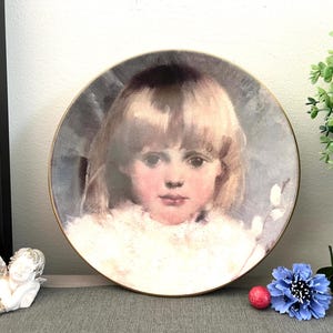 Puede incluir: Un plato decorativo con un retrato de un niño pequeño con cabello rubio y un cuello con ribete de piel blanca. El plato tiene un borde dorado y está sobre un fondo suave y apagado. Hay objetos decorativos en primer plano.