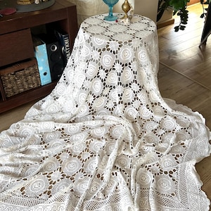 May include: A white circular crochet tablecloth featuring an intricate pattern of repeating circular motifs and openwork mesh detailing. The fabric drapes over a round table, extending down to the floor with a scalloped, decorative border edge.