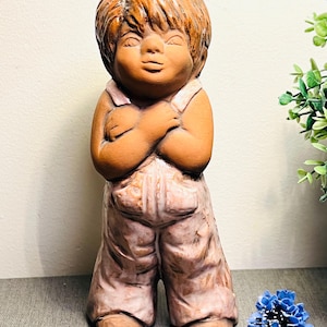 Könnte beinhalten: Terrakotta-Figur eines Kindes mit verschränkten Armen, das eine Latzhose trägt. Die Statue hat einen hellbraunen Teint und hellbraunes Haar. Die Latzhose hat eine verblasste rosa Farbe. Die Figur ist ein Dekorationsartikel, geeignet für die Innenausstellung.