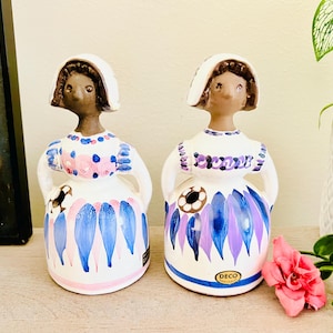 May include: Two ceramic figurines of women wearing white bonnets and colourful dresses with blue and pink floral patterns. The figurines are standing on a wooden surface. The figurines are labelled "DECO" on the bottom.