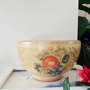 Puede incluir: Un cuenco de cerámica de color crema con un diseño pintado a mano de frutas y vides. El cuenco tiene un esmalte moteado y una forma redondeada.
