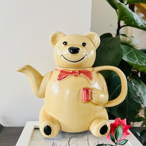 Puede incluir: Una tetera de cerámica con forma de oso marrón claro con una pajarita roja. La tetera tiene un pico y una asa. El oso tiene ojos negros y una nariz negra.