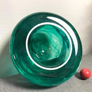 Peut inclure: Un bol rond en verre vert émeraude avec un anneau intérieur blanc. Le bol présente un motif tourbillonnant et marbré. Un petit objet rond rouge est à côté du bol. C'est un objet décoratif.