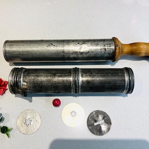 May include: Two vintage metal cookie presses with wooden handles. The presses are silver in color and have a star-shaped design. There are three cookie cutters on a white surface, one with a star shape, one with a flower shape, and one with a wavy design.