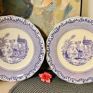 Może przedstawiać: Dwa vintage talerze porcelanowe w kolorze niebieskim i białym z ozdobną obwódką i centralnym wzorem przedstawiającym niebiesko-białą scenę z palmami i budynkiem.