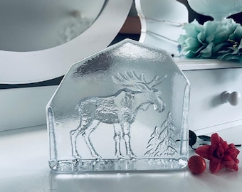 May include: A clear glass paperweight featuring an etched design of a moose standing in a forest. The moose is facing right and has a detailed, textured design. The forest is represented by two trees.