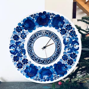 Könnte beinhalten: Eine weiße Keramikuhr mit einem blauen und weißen Blumendesign. Das Zifferblatt der Uhr ist weiß mit schwarzen römischen Ziffern als Stundenmarkierungen und schwarzen Zeigern. Die Uhr hat einen Durchmesser von 30 cm.