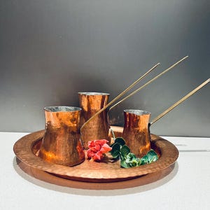 May include: Three copper coffee pots with long handles on a hammered copper tray with a small bouquet of pink flowers.