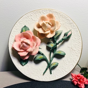 Puede incluir: Un plato de cerámica blanco con un borde dorado presenta un diseño 3D de dos rosas rosas y melocotón con hojas verdes. Las rosas están en relieve y parecen esculpidas.