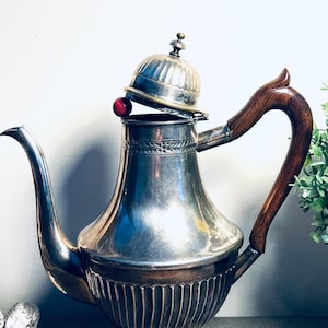 May include: A silver teapot with a curved spout and a wooden handle. The lid is slightly ajar, revealing a red sphere. The teapot has a decorative, ribbed base and a detailed lid. A vintage tea or coffee pot.