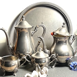May include: A silver tea set with ornate detailing, including a teapot, creamer, sugar bowl, and a serving tray. The set has a classic design with curved handles and decorative feet. The background includes a silver tray and a few decorative flowers.