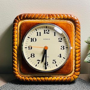 May include: A square, vintage Kienzle quartz clock with a cream-coloured face and black numerals. The clock has a brown frame with a decorative rope-like border. The hands are black and orange. The clock is approximately 20 cm square.