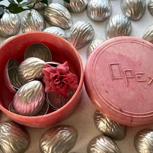 May include: A round pink container with a matching lid embossed with the text Opex, filled with several silver-colored metal walnut-shaped molds and a single pink fabric flower. Additional silver metal walnut molds are scattered around the container.