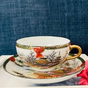 May include: A white teacup and saucer set with a gold rim and handle. The cup and saucer feature a detailed landscape design with red, gold, and black accents. The set is placed on a white surface.