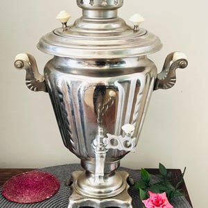 May include: A metallic silver samovar featuring a fluted body, two side handles with cream-colored accents, a matching lid with two small knobs, and a central spigot with a cream-colored handle. The base is square with four decorative feet.