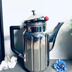 May include: A vintage silver-plated coffee pot with a black handle and a lid with a red knob. The pot has a ribbed design and a long spout. The background includes a small cherub statue and a blue flower.