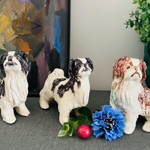 Könnte beinhalten: Drei Keramik-Hundefiguren in Weiß, Schwarz und Braun. Die Hunde sind in verschiedenen Posen dargestellt. Eine blaue Blume und eine rote Beere befinden sich vor den Hunden. Eine grüne Pflanze steht in einer türkisfarbenen Vase im Hintergrund.