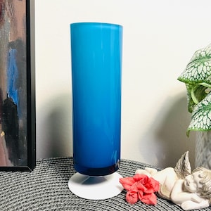 Puede incluir: Un jarrón de vidrio azul alto con una base blanca. El jarrón está esmerilado y tiene un degradado de color de azul oscuro en la parte inferior a azul claro en la parte superior.