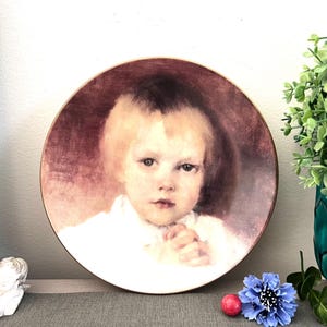 Puede incluir: Un plato decorativo con un retrato de un niño con cabello rubio y un atuendo blanco. El plato tiene un fondo granate y un borde dorado. El plato se exhibe con un jarrón turquesa y una flor azul.