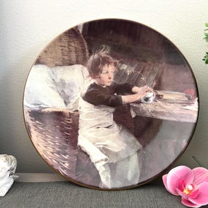 Puede incluir: Plato decorativo redondo con una pintura vintage de un niño en una mesa. El niño lleva una camisa oscura y un delantal blanco. El plato tiene un borde dorado y se exhibe sobre una superficie gris.