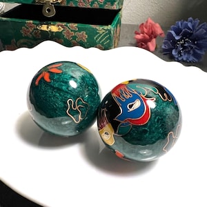 Può includere: Due sfere decorative in cloisonné verde con disegni floreali e di uccelli colorati. Le sfere hanno una finitura lucida e sono impreziosite da metallo color oro. Le sfere sono esposte su un piatto bianco smerlato.