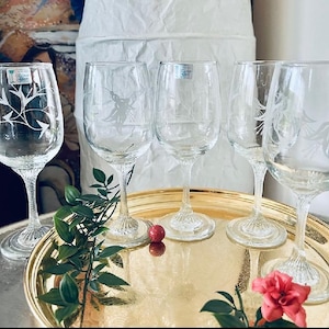 May include: Set of four clear glass wine glasses with etched floral designs. The glasses are arranged on a gold tray with a green plant and a red berry.