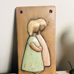 May include: A rectangular ceramic wall plaque with a relief sculpture of two figures embracing. The figures have blonde hair and wear a light green and pink dress. The plaque is light brown with two holes at the top.