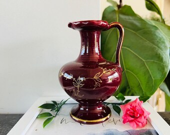 May include: A small, burgundy ceramic vase with gold floral accents. The vase has a wide mouth and a handle.