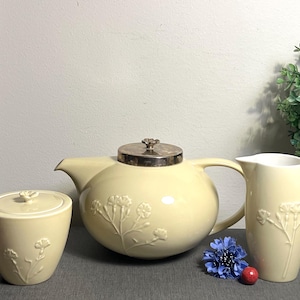May include: A cream-colored tea set with floral embossed detailing. The set includes a teapot with a silver lid, a sugar bowl with a lid, and a creamer. A blue flower and a red berry are next to the set.