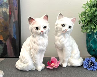 May include: Two white ceramic cat ornaments with detailed features, including pink ears and noses, and expressive eyes. The cats are seated upright, with a fluffy texture. A pink orchid and blue flower are in front of the cats.