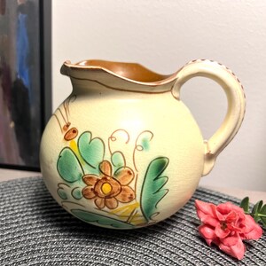 May include: A cream-colored ceramic pitcher with a brown handle and a floral design in shades of green, brown, and yellow.