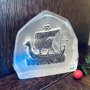 May include: A clear glass paperweight featuring a silver Viking ship design. The ship has a sail and a dragon head on the bow. The paperweight has a small blue and yellow sticker on the side.