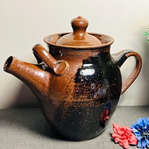 May include: A handcrafted, brown ceramic teapot with a unique spout and handle design. The teapot features a lid and a dark, speckled glaze, with a capacity of approximately 1.5 liters. The teapot is a rustic, vintage style.