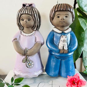 Könnte beinhalten: Zwei Keramikfiguren, ein Mädchen und ein Junge. Das Mädchen trägt ein lavendelfarbenes Kleid und hält eine Blume. Der Junge trägt ein blaues Hemd und eine blaue Hose und hält einen kleinen weißen Hund.