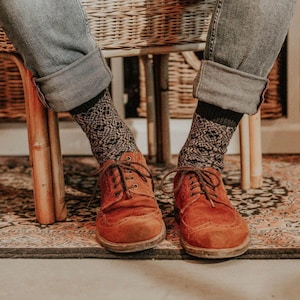 May include: A pair of brown leather lace-up shoes with a patterned design on the socks. The shoes are worn by a person sitting on a wicker chair.