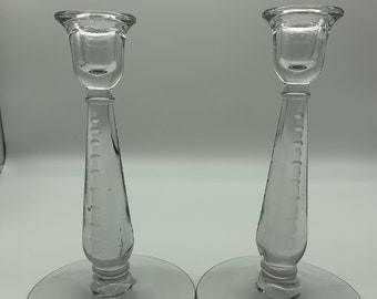 May include: A pair of clear glass candlesticks. Each has a circular base, a long, slender stem with etched detailing, and a cup-shaped top for a candle. The candlesticks are identical in design and size.