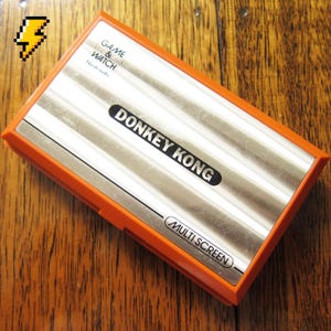 May include: A rectangular orange and silver Nintendo Game & Watch handheld electronic game. The silver faceplate features the text GAME & WATCH, Nintendo, DONKEY KONG, and MULTI SCREEN. A small pixelated yellow lightning bolt icon is in the corner.