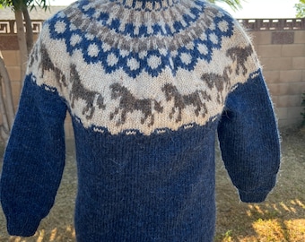 May include: A blue, beige, and gray knitted sweater with a high collar. The sweater features a pattern of horses and geometric shapes around the yoke. The sleeves and body are a solid blue color, and the sweater is displayed on a mannequin.