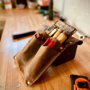 May include: A brown leather tool belt with various tools, including chisels and a tape measure. The belt has multiple pockets and is stitched with red thread. The tools are visible inside the pockets, and the tape measure is orange and black.