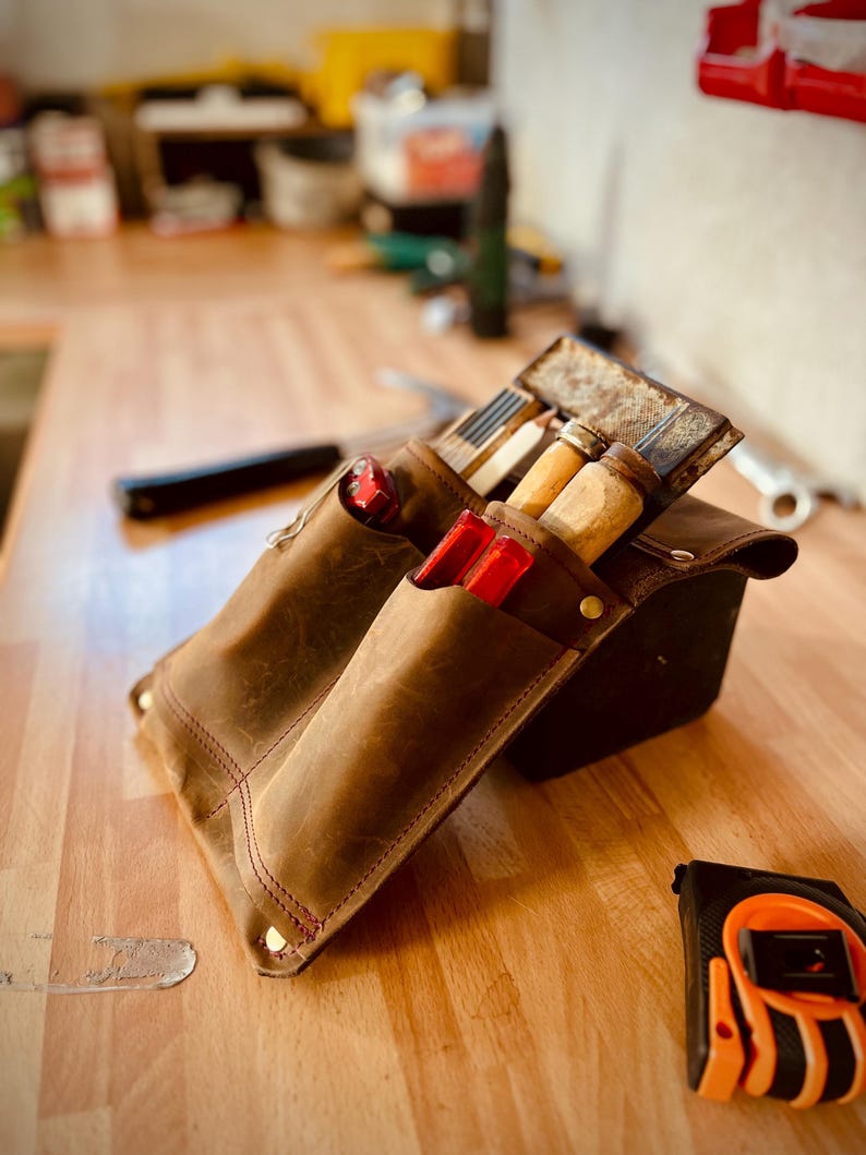 May include: A brown leather tool belt with various tools, including chisels and a tape measure. The belt has multiple pockets and is stitched with red thread. The tools are visible inside the pockets, and the tape measure is orange and black.