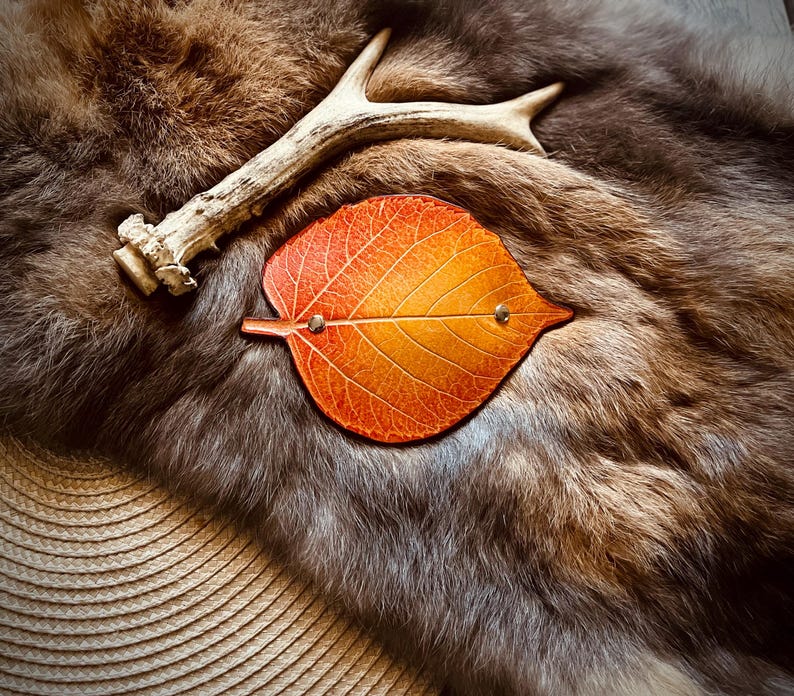 May include: An orange and yellow leaf-shaped leather coaster with two small metal rivets. The coaster is set on a backdrop of brown fur and a deer antler. The overall theme is rustic and natural.
