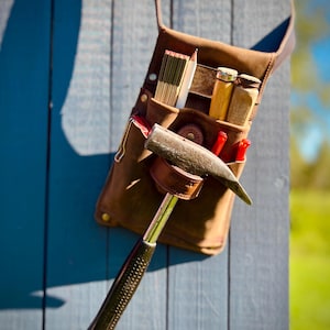 May include: A brown leather tool belt with various carpentry tools, including a hammer, pencils, and a folding ruler. The tools are arranged in pockets and loops. The belt is hanging on a blue wooden surface.