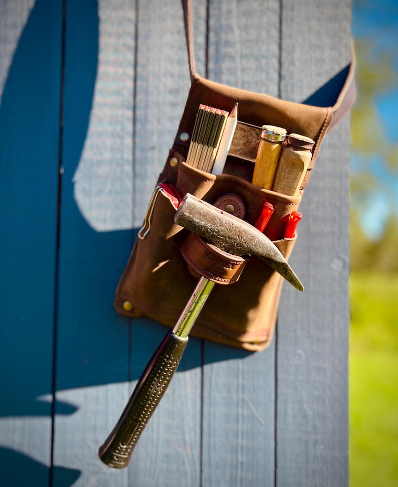 May include: A brown leather tool belt with various carpentry tools, including a hammer, pencils, and a folding ruler. The tools are arranged in pockets and loops. The belt is hanging on a blue wooden surface.