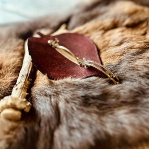 May include: A burgundy leather item with a gold-colored metal hair clip. The clip is decorated with small, clear stones. The items are displayed on a textured, brown and gray fur-like surface, with a piece of antler in the background.