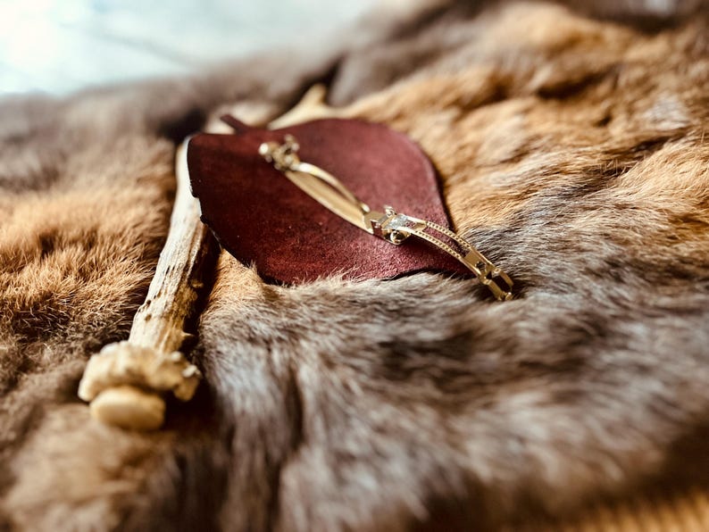 May include: A burgundy leather item with a gold-colored metal hair clip. The clip is decorated with small, clear stones. The items are displayed on a textured, brown and gray fur-like surface, with a piece of antler in the background.