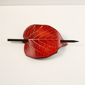 May include: A red leaf-shaped hair barrette with a dark wooden stick. The barrette has a detailed vein pattern and a smooth, polished finish. The hair accessory is designed to hold hair in place.